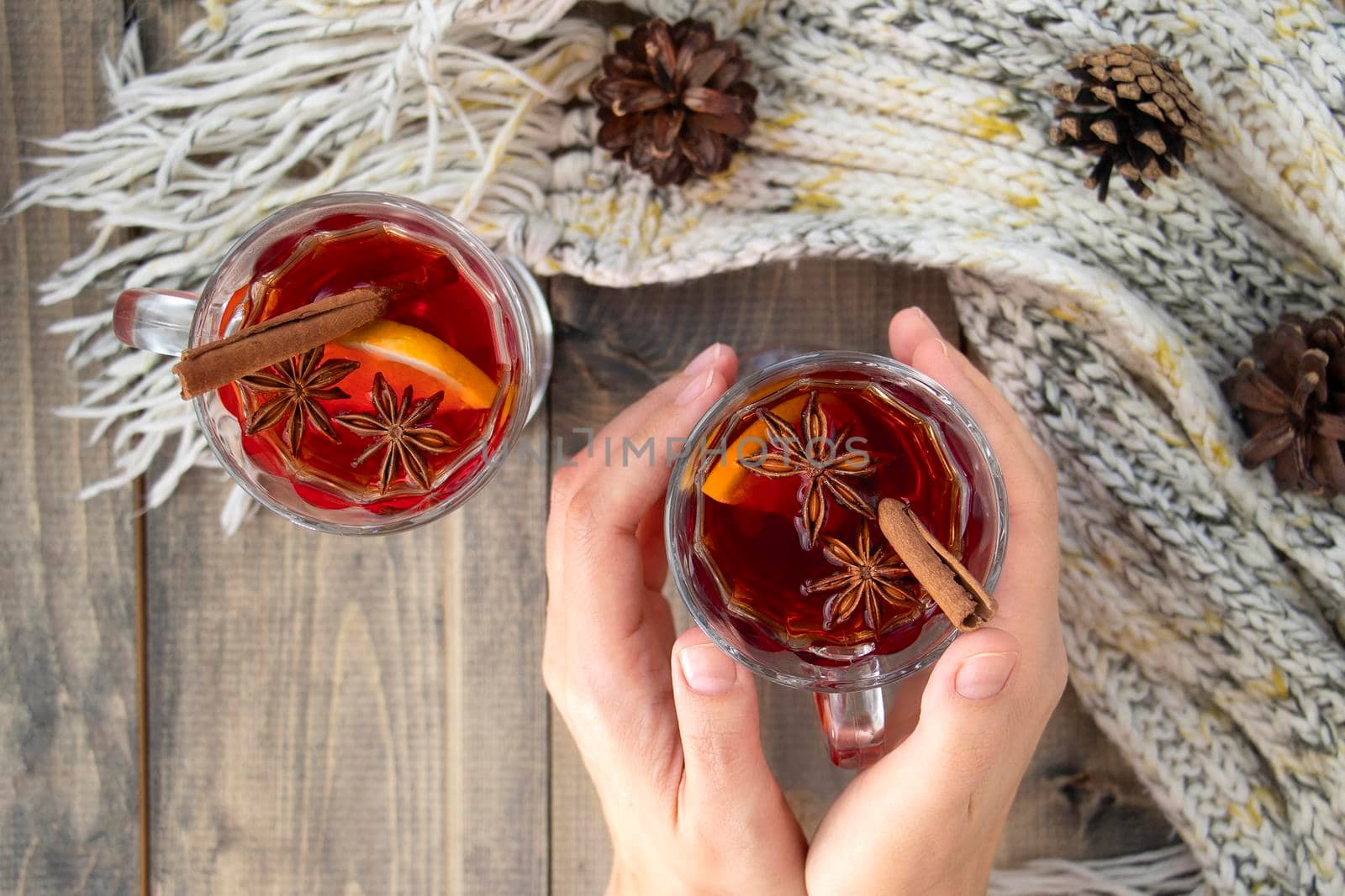 female hands hold a glass of hot punch. mulled wine on a wooden table next to a warm knitted scarf. a warming drink in winter or autumn. Soft focus. view from above. flat lay