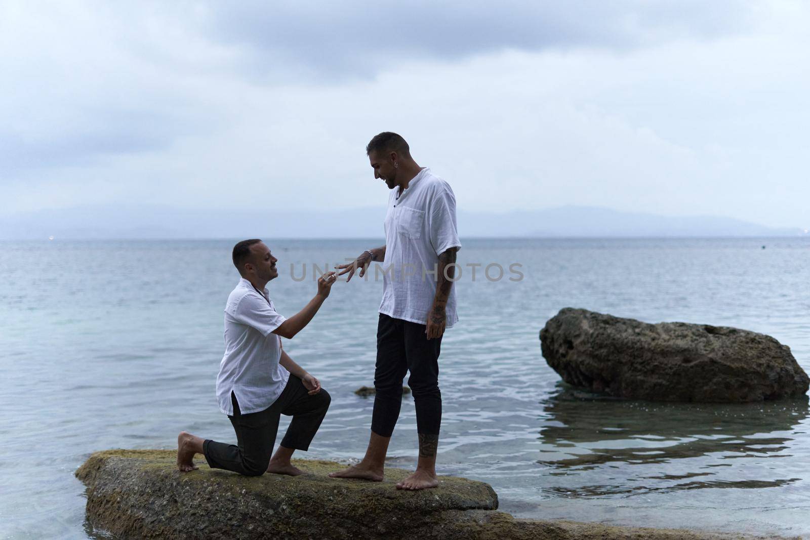 Gay man on his knees asking his partner to marry him in the middle of rocks in the sea
