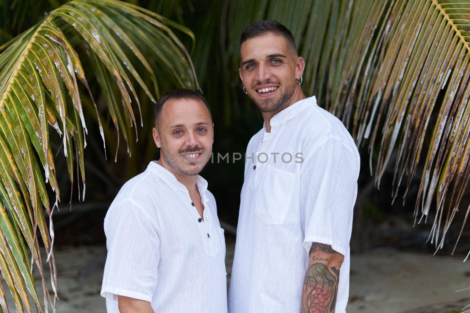 Portrait of two gay men looking to the camera with happy and friendly expression next to a palm tree