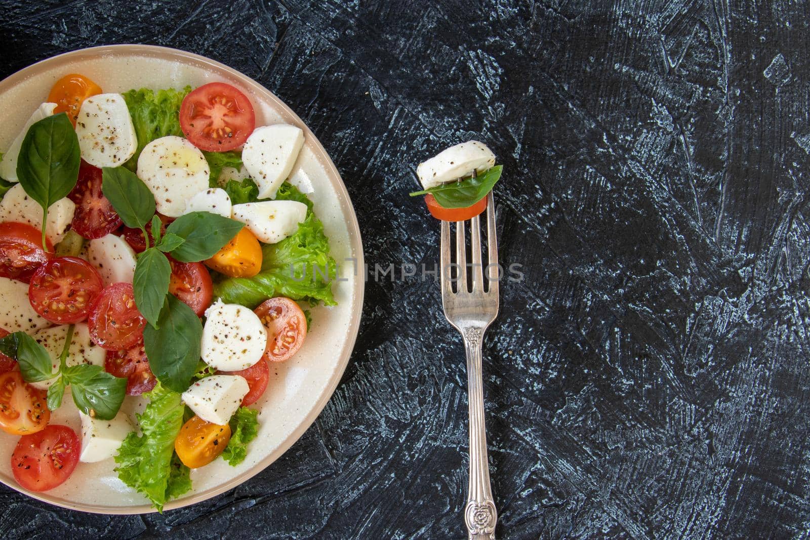 flat lay with fork and plate of vegetable salad with cheese. serving Caprese salad with tomatoes, mozzarella, basil and olive oil with copy space on black texture background. top view. Soft focus