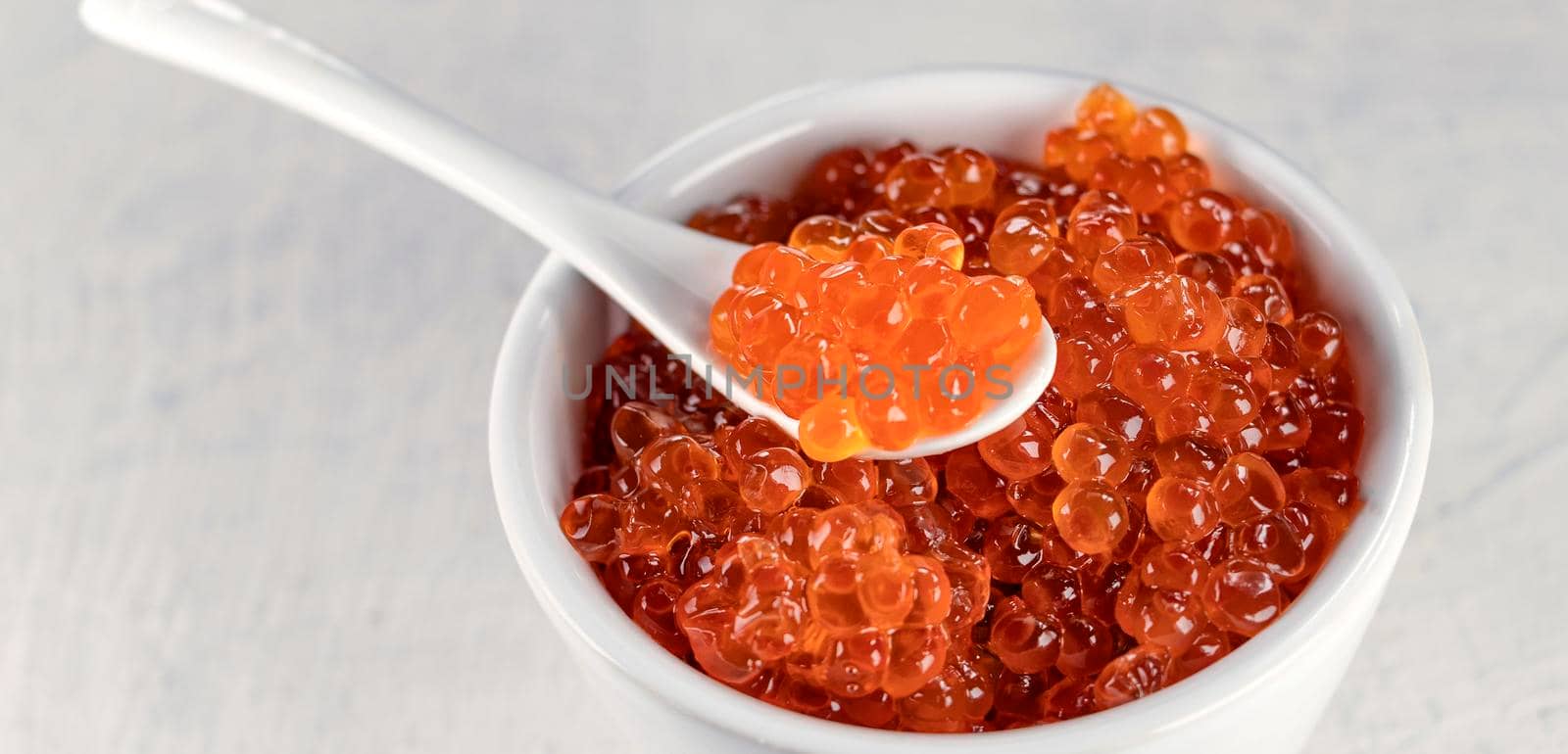 banner with close up delicious, crumbly, large red caviar in a white cup with a spoon on a white background. by Leoschka