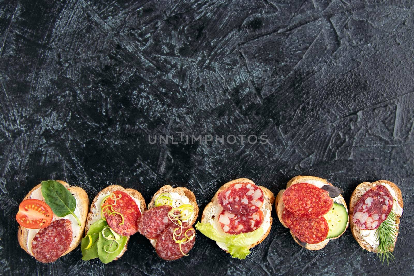 flat lay with sandwiches with sausage, cream cheese, tomatoes, dill, cucumbers, and basil. serving a snack for appetizers on black background with copy space. by Leoschka