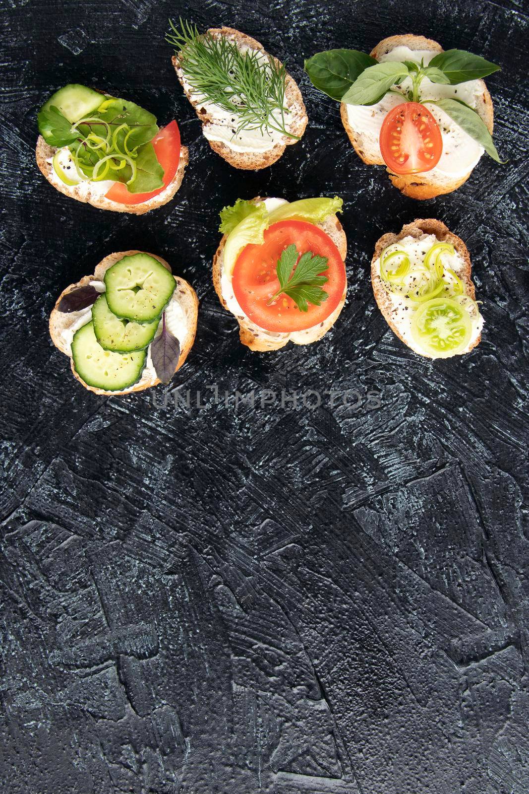 vertical photo with serving vegetarian sandwiches for appetizers on black textured background. various vegetable sandwiches with cream cheese, tomatoes, dill, cucumbers, leeks and basil. copy space