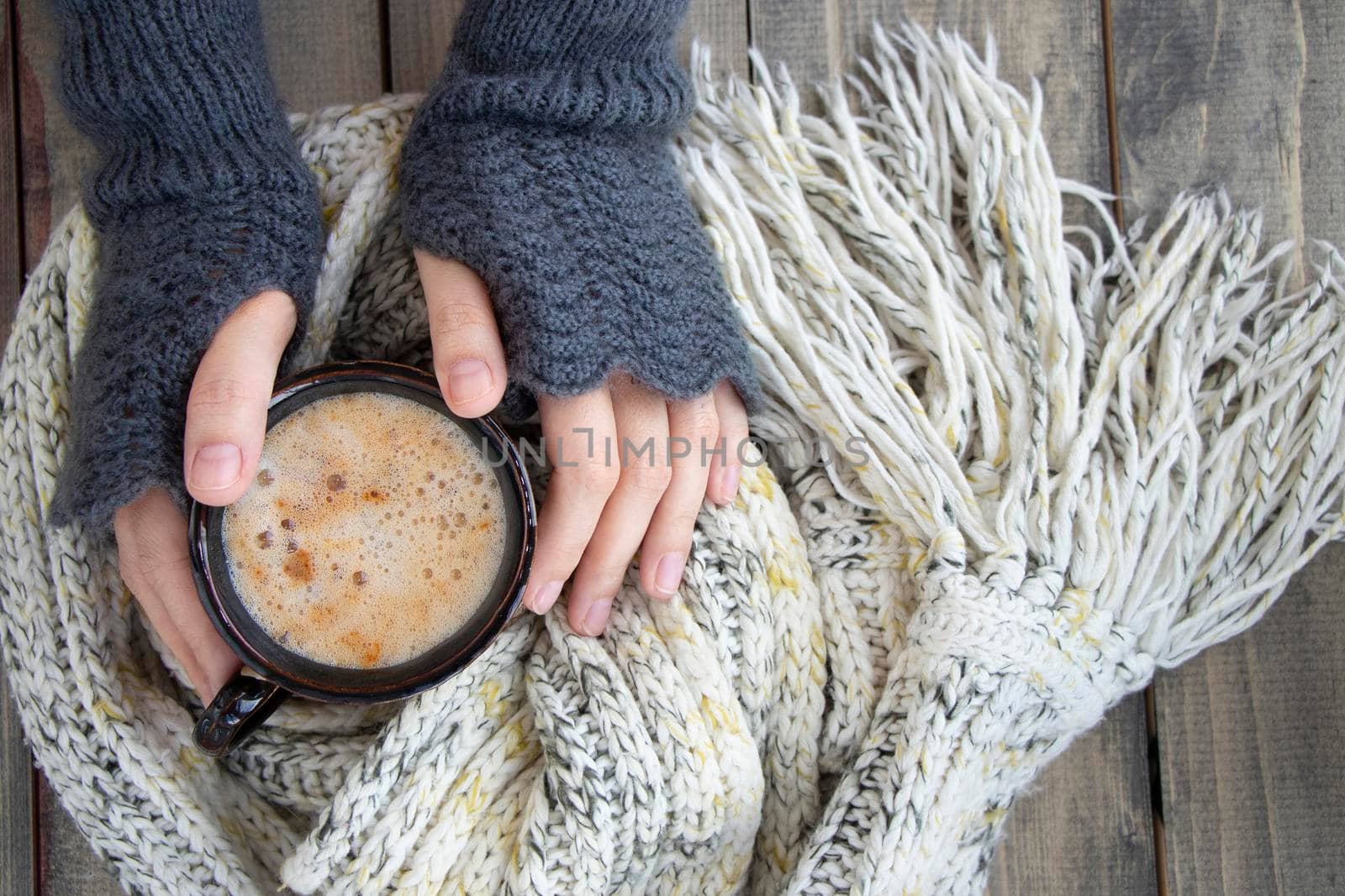 female hands in mittens hold a cup of hot drink with foam. mug of coffee on a wooden table next to a warm knitted scarf. Soft focus. by Leoschka