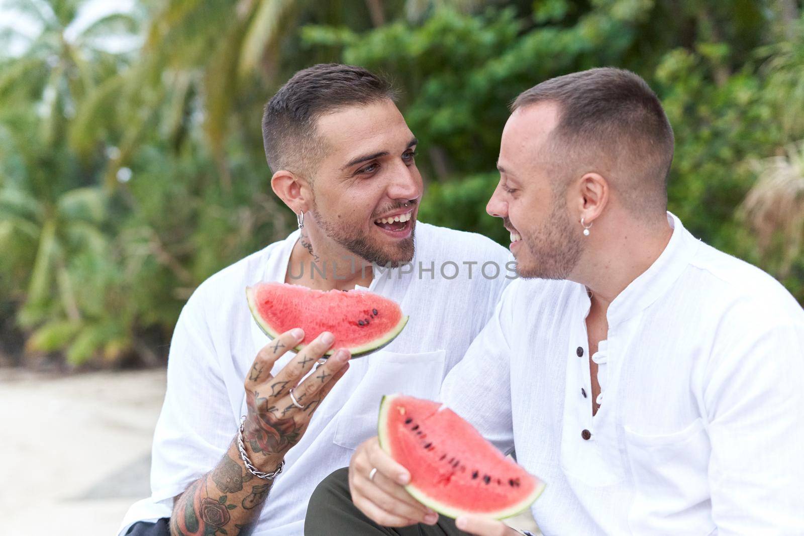 Happy homosexual couple chatting distracted while eating watermelon