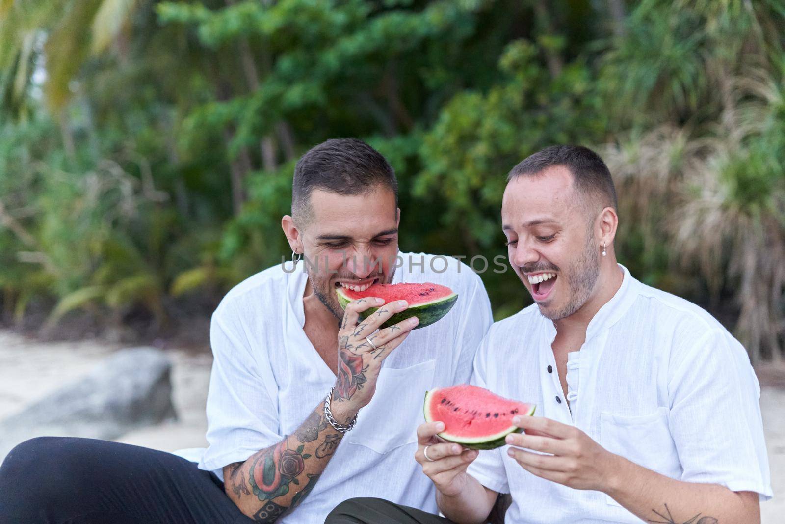 Smiling gay couple eating watermelon and chatting relaxed sitting on a tropical beach in Thailand
