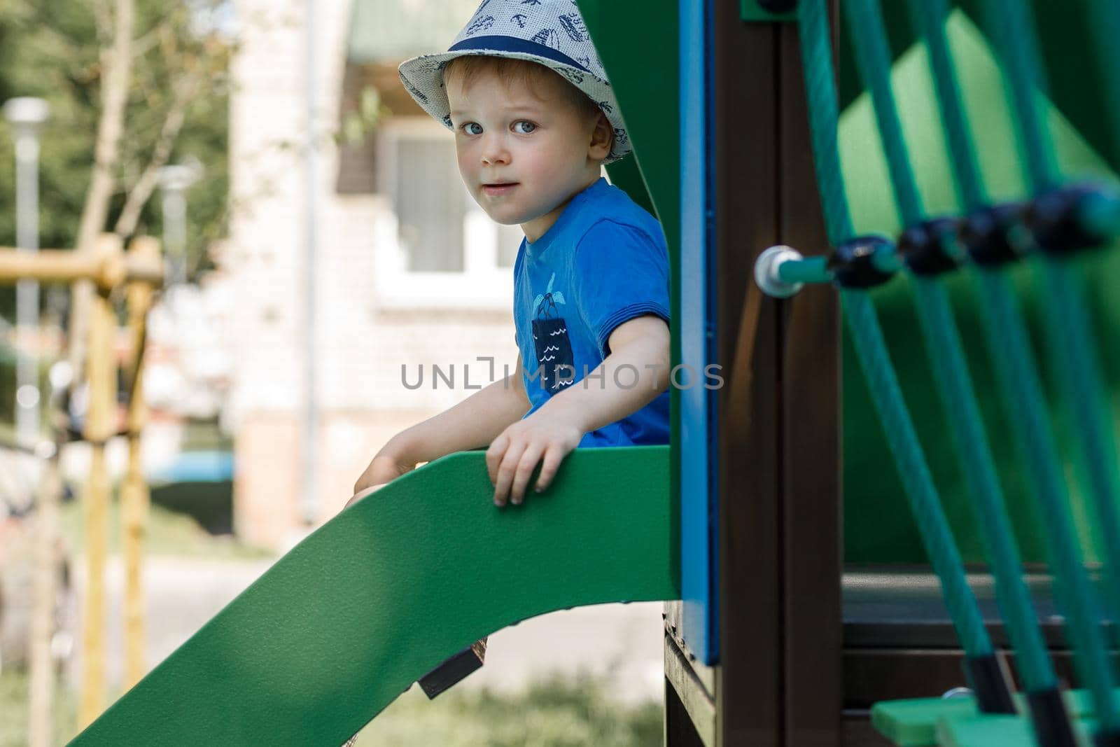 Cheerful little boy in a blue T-shirt and beige shorts rolls down a metal slide in city park by Lincikas