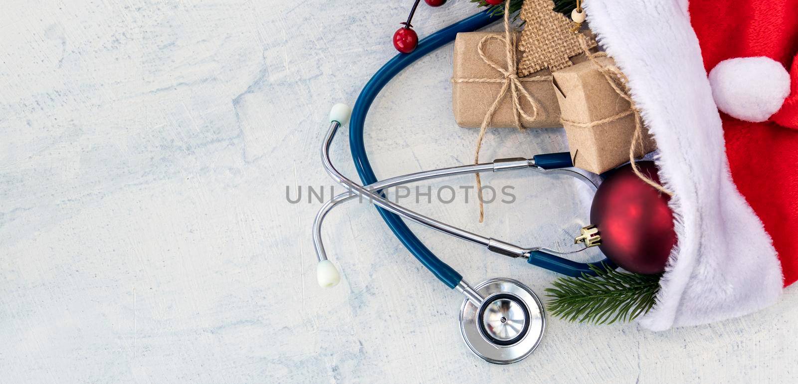 banner with Santa Claus hat and stethoscope, Christmas gifts, and a branch of Christmas tree with red Christmas ball on a white concrete table. Xmas. Copy space. by Leoschka