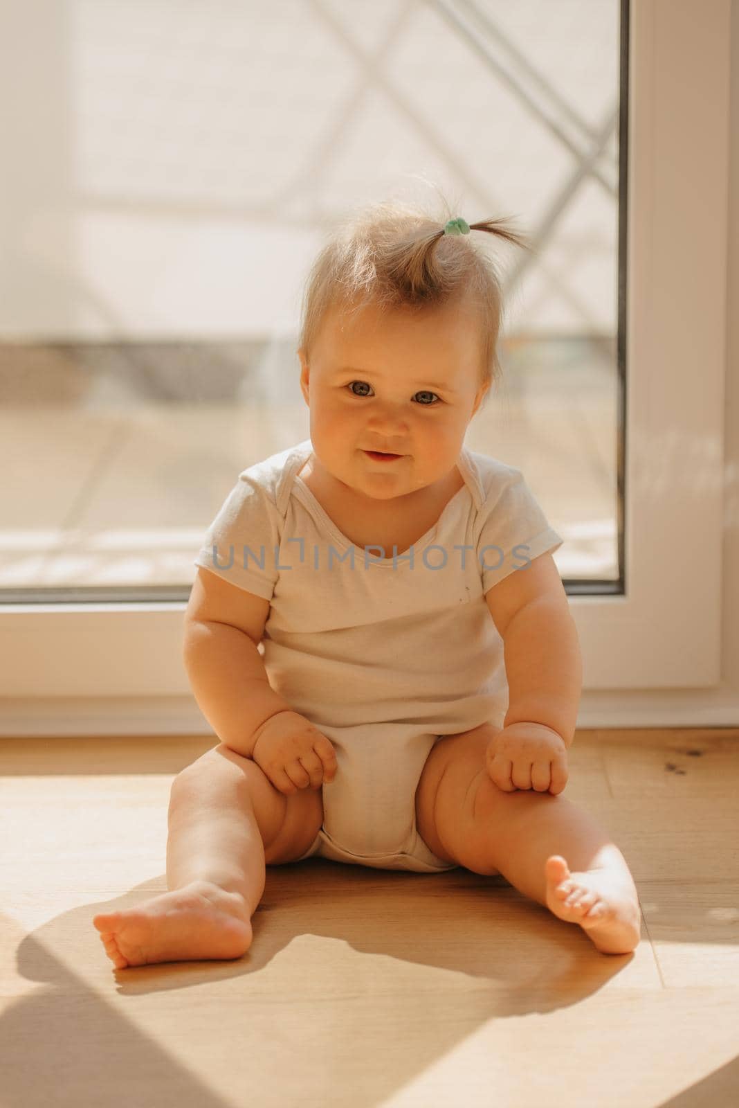 A 7-month girl is sitting near a balcony door in a bodysuit at home. A cute infant is waiting for her parents in the sunlight.