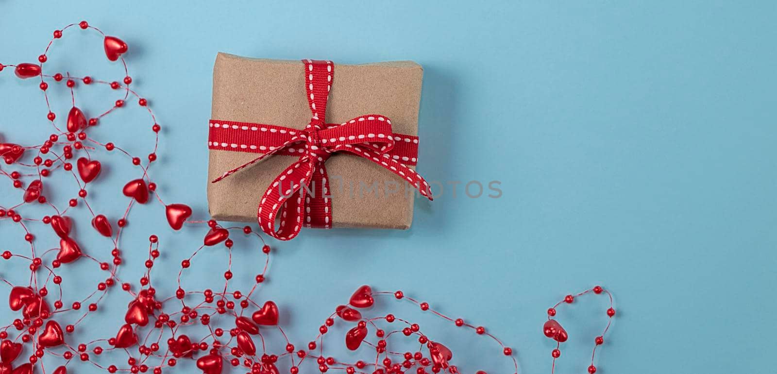 banner with concept of minimalism valentine's day. gift with red ribbon on pastel blue background with red beads with hearts with copy space. Soft focus. flat lay
