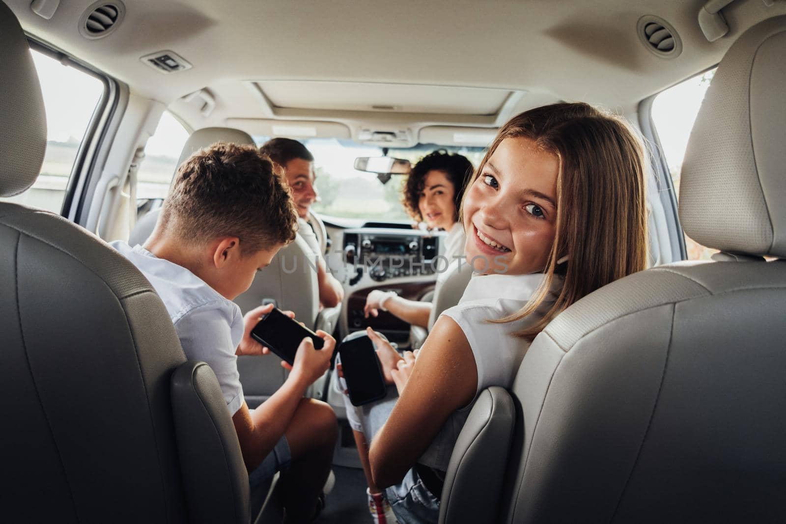 Cheerful Caucasian Teenage Girl Smiles Into Camera While Sitting in Minivan Car with Her Brother, Mother and Father, Happy Four Members Family Enjoying Weekend Road Trip