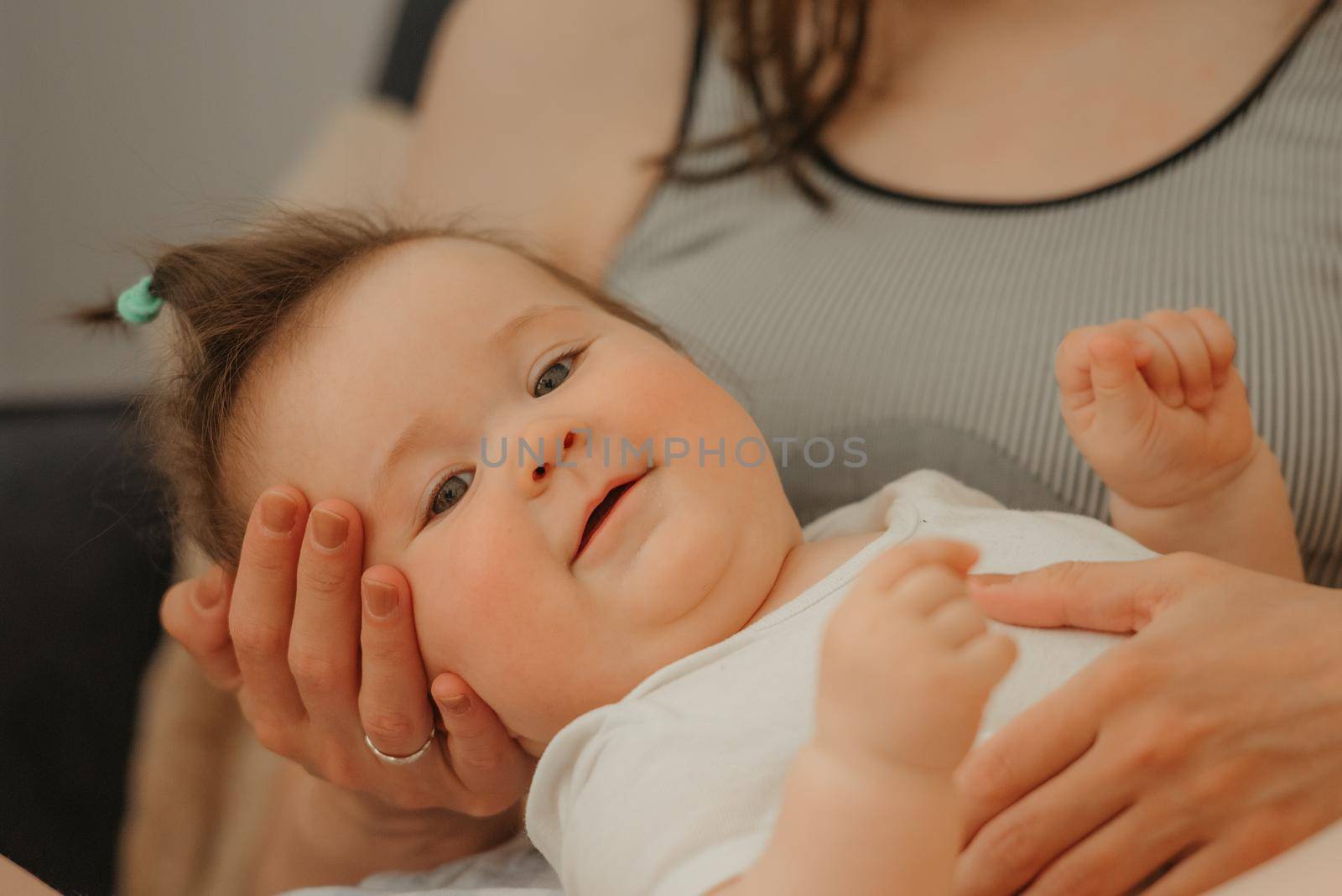 A close portrait of a 7-month girl in the arms of her mother who is smiling at home. A curious infant in a bodysuit is lying on the knees of her mother.