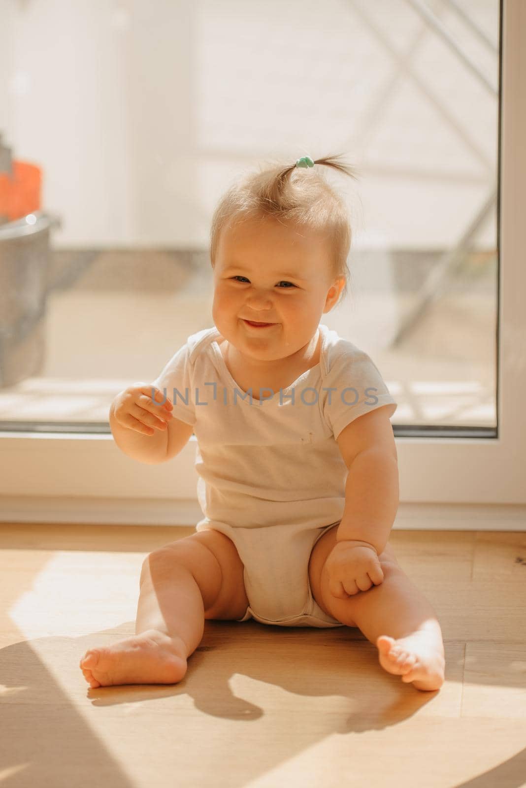 A kind 7-month girl is sitting near a balcony door in a bodysuit at home. A cute infant is laughing in the sunlight.