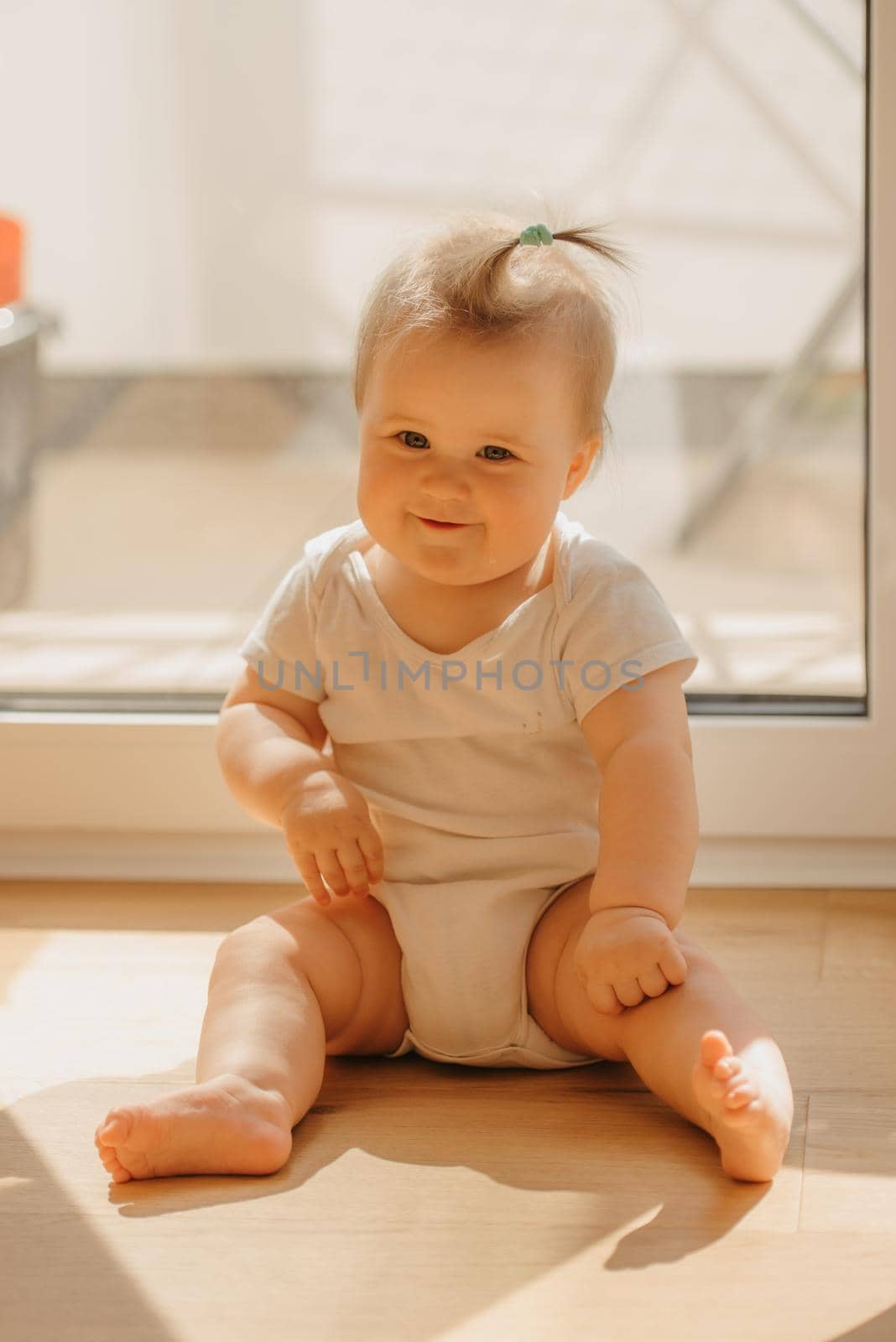 A kind 7-month girl is sitting near a balcony door in a bodysuit at home. A cute infant is smiling in the sunlight.