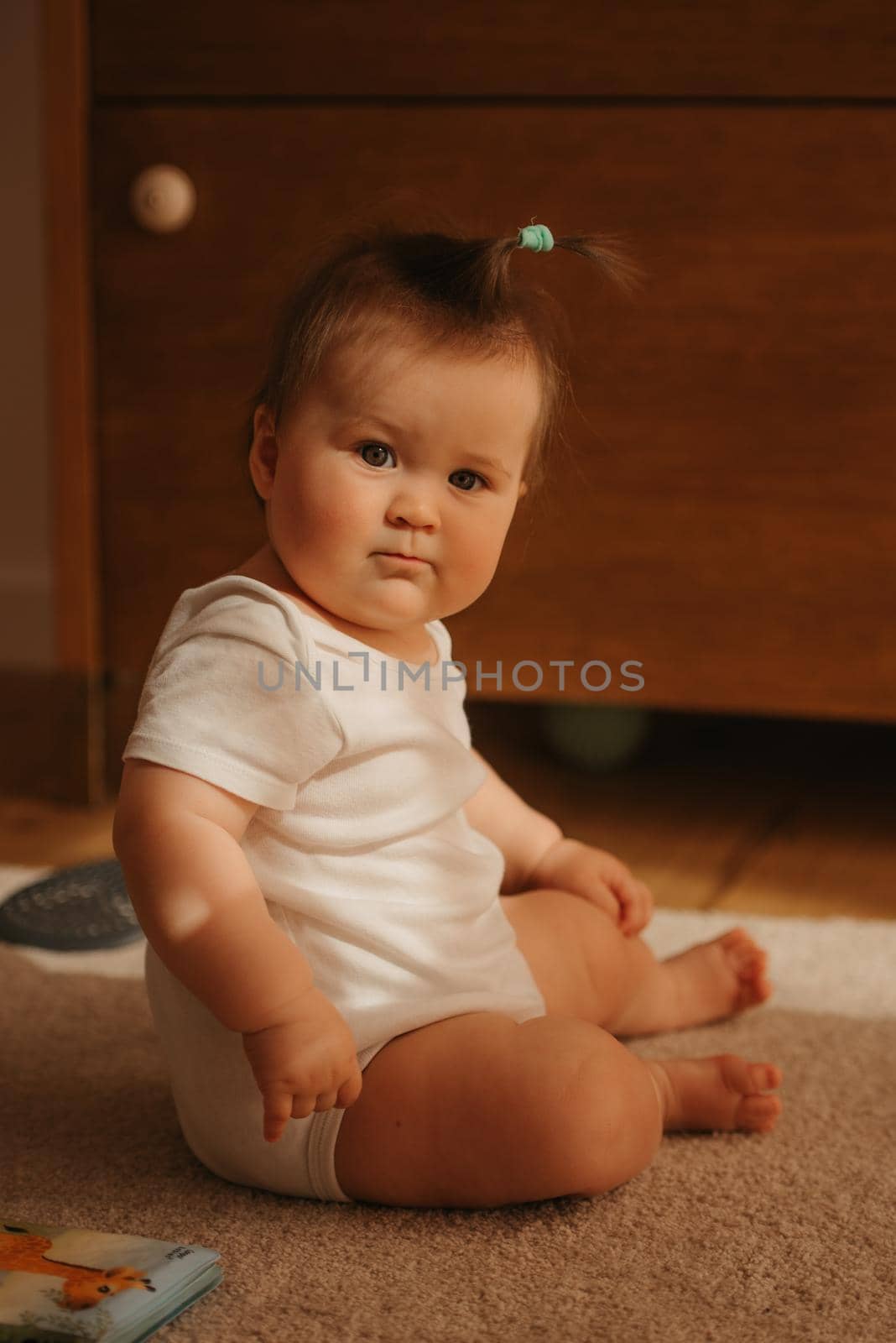 A 7-month girl is sitting near a dresser in a bodysuit at home. A serious infant in the sunlight.