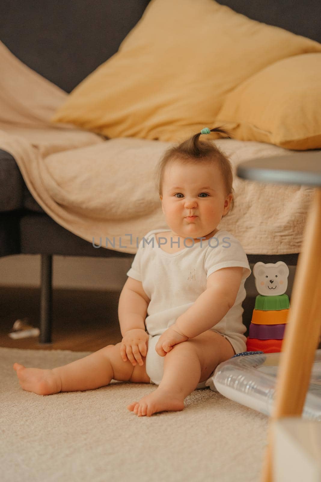 A 7-month girl is sitting near a sofa and making faces in a bodysuit at home. A smiling infant in the sunlight.