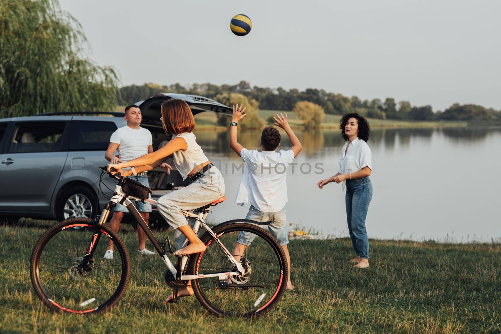 Teenage Girl Riding Bicycle While Mother and Father Playing Volleyball with Her Brother Outdoors by Lake, Happy Four Members Family Enjoying Weekend Road Trip on Minivan Car