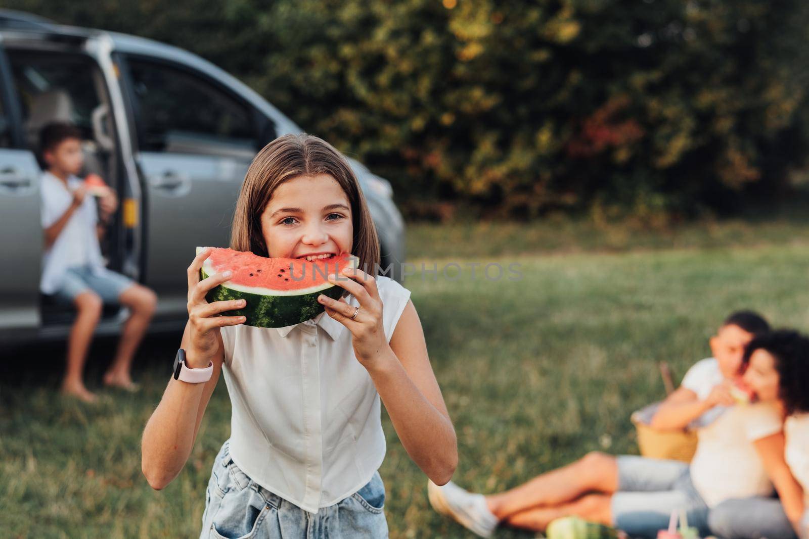 Caucasian Teenage Girl Eating Watermelon Outdoors, Family on Background Having Picnic While Traveling by Minivan Car