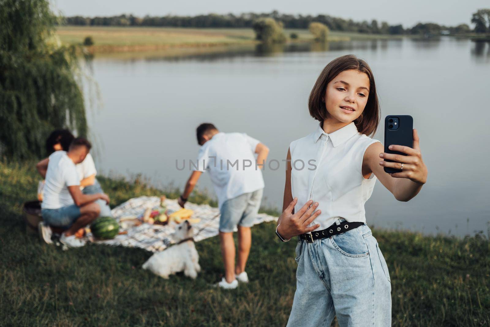 Teenage Caucasian Girl Making Selfie on Smartphone While Her Family on the Background Having Picnic by Lake Outdoors