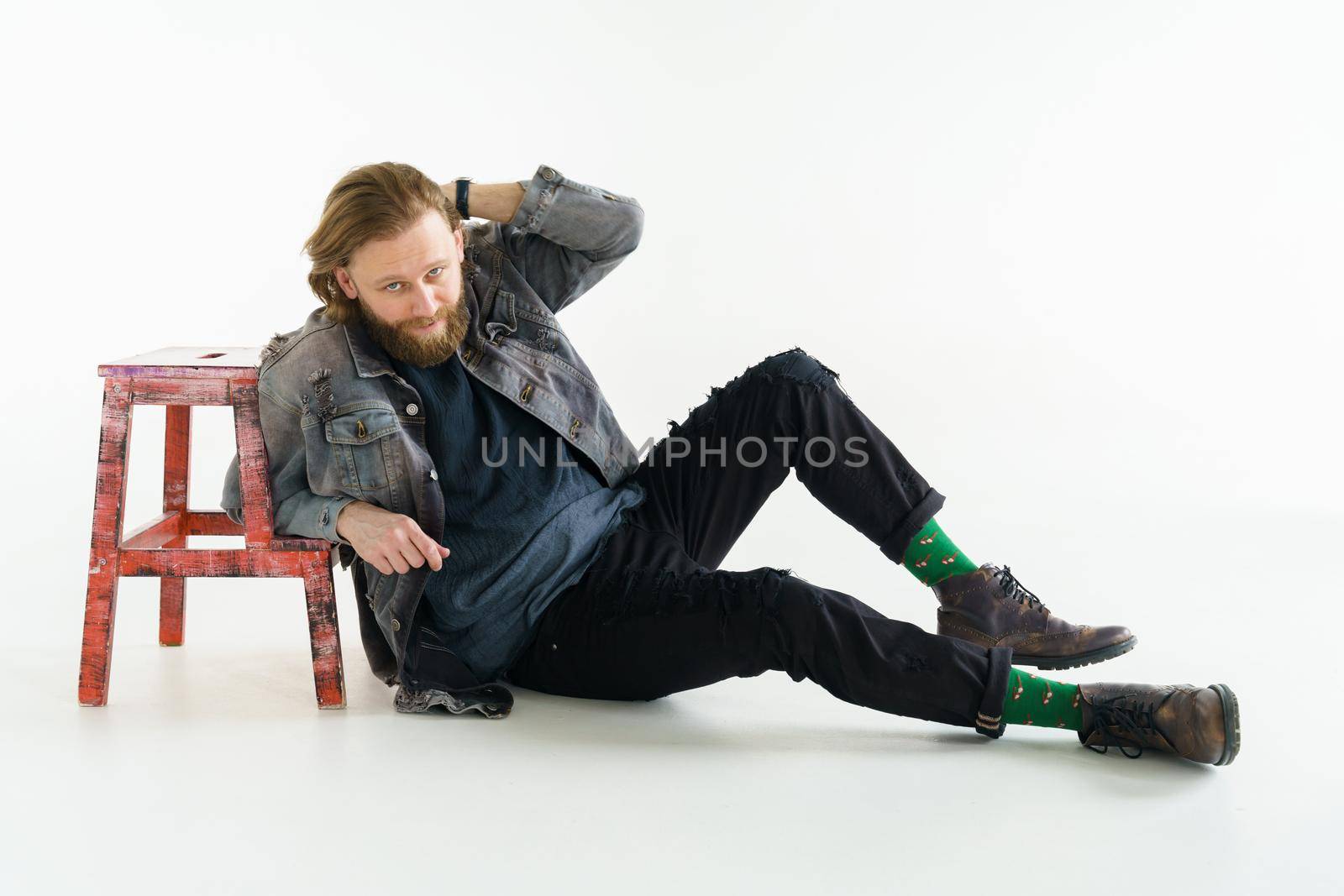 a handsome brutal bearded and long-haired man sits on the floor leaning on a chair on a white background, he is dressed in a denim jacket and pants with bright green socks, dressed like a hipster. High quality photo