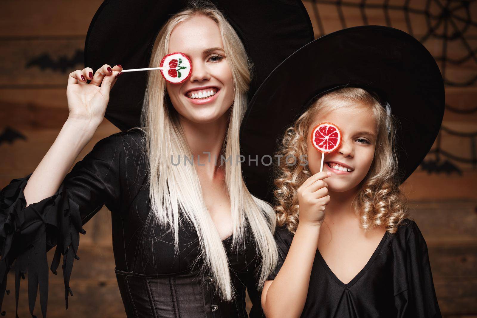 Halloween Concept: beautiful caucasian mother and her daughter in witch costumes celebrating Halloween with Halloween candy and sweet over bats and spider web on Wooden studio background