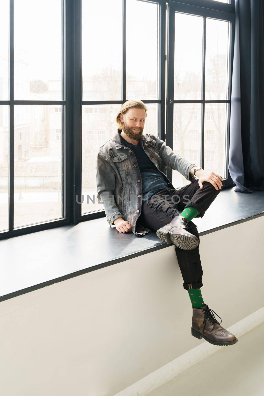 a handsome brutal bearded and long-haired man sits on the windowsill of a huge window, he is dressed in a denim jacket and pants with bright green socks, dressed like a hipster. High quality photo