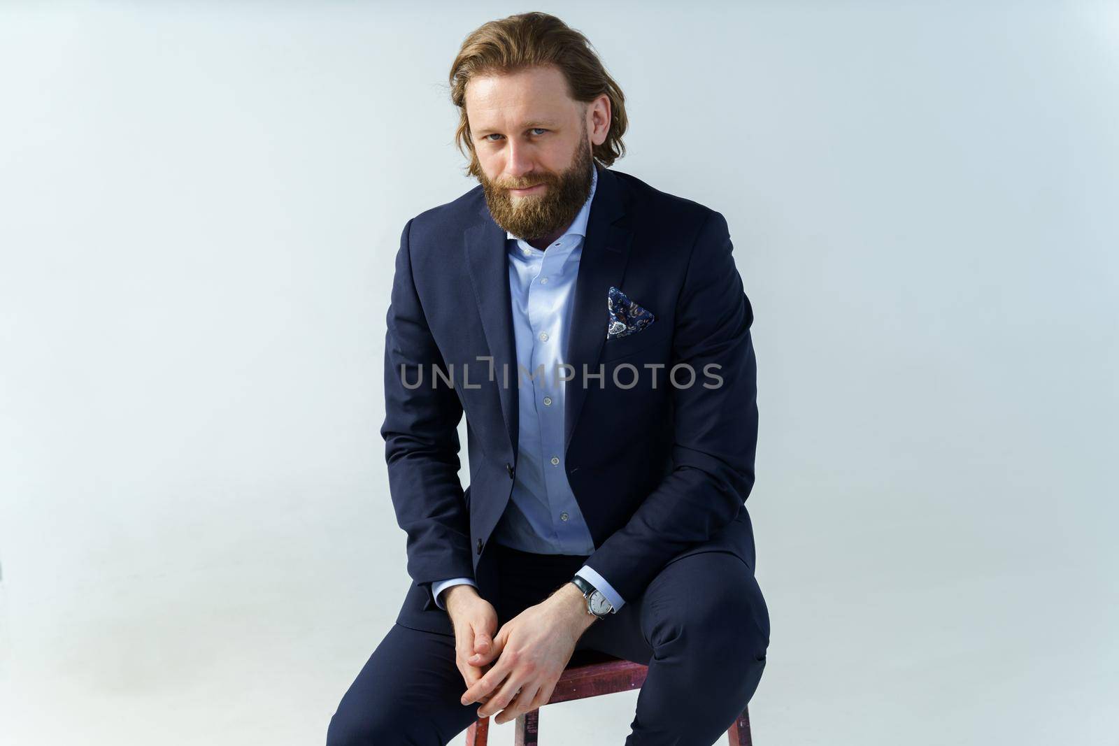 a handsome brutal bearded and long-haired man, a stylish businessman sitting on a chair with a white background. High quality photo