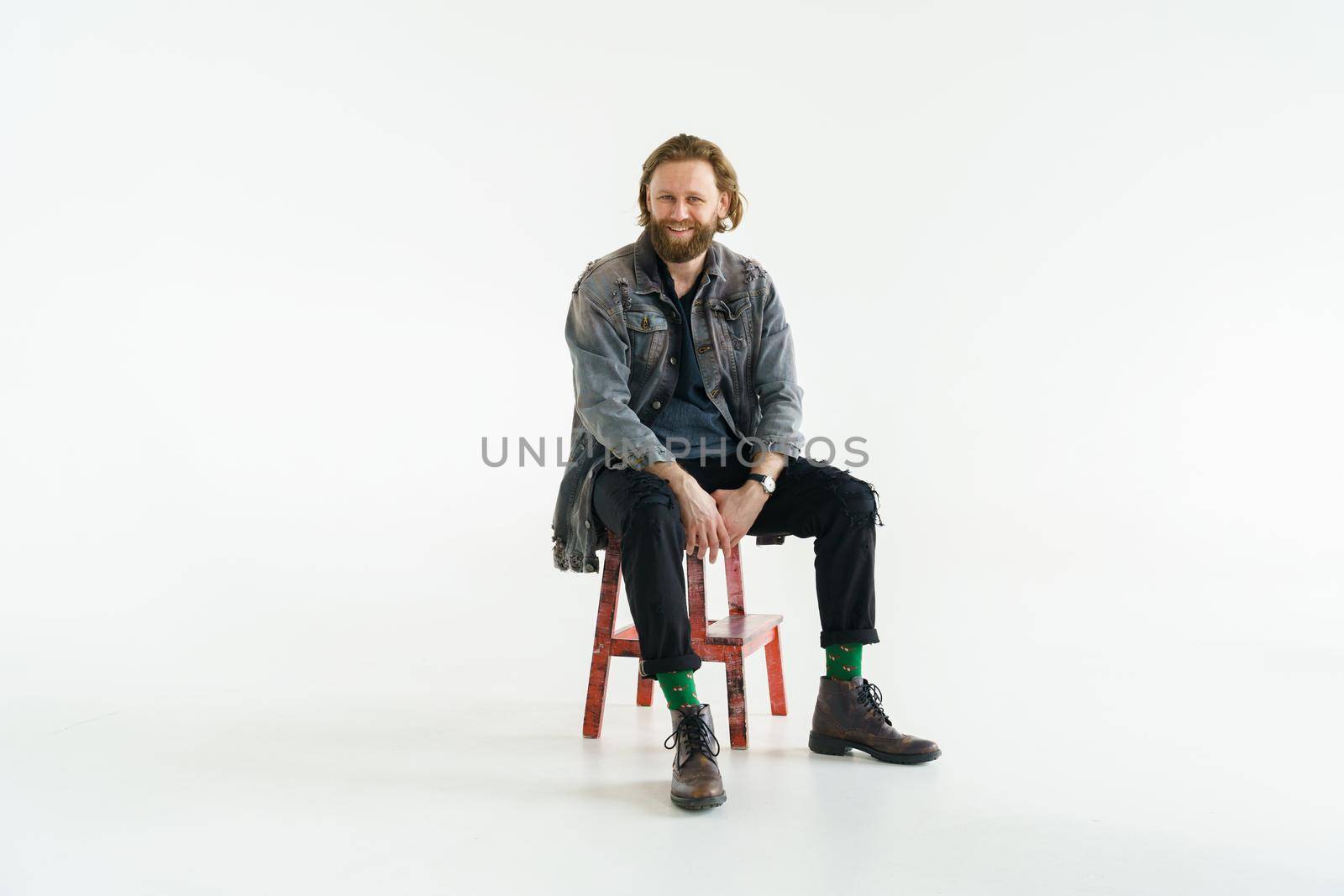 a handsome brutal bearded and long-haired man is sitting on chair on a white background, he is dressed in a denim jacket and pants with bright green socks, dressed like a hipster. High quality photo