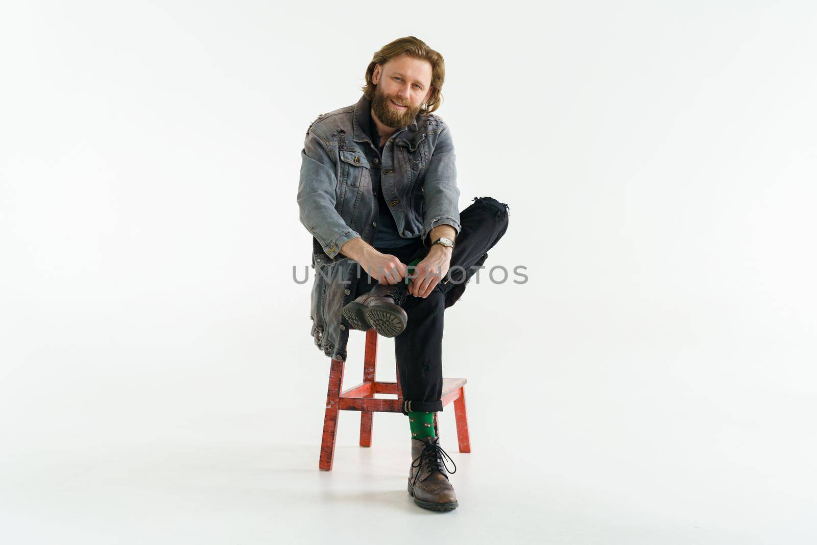 a handsome brutal bearded and long-haired man is sitting on chair on a white background, he is dressed in a denim jacket and pants with bright green socks, dressed like a hipster. High quality photo