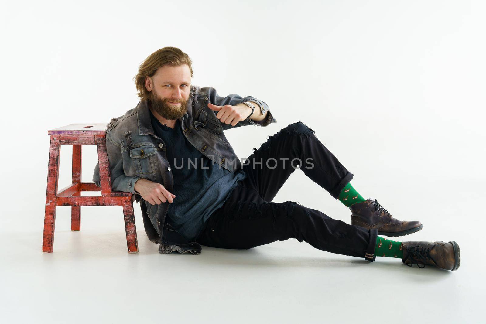a handsome brutal bearded and long-haired man sits on the floor leaning on a chair on a white background, he is dressed in a denim jacket and pants with bright green socks, dressed like a hipster. High quality photo