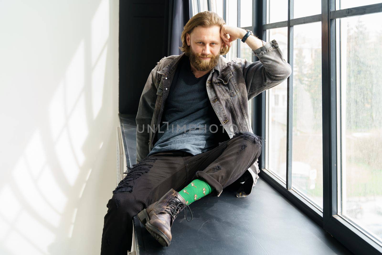 a handsome brutal bearded and long-haired man sits on the windowsill of a huge window, he is dressed in a denim jacket and pants with bright green socks, dressed like a hipster. High quality photo