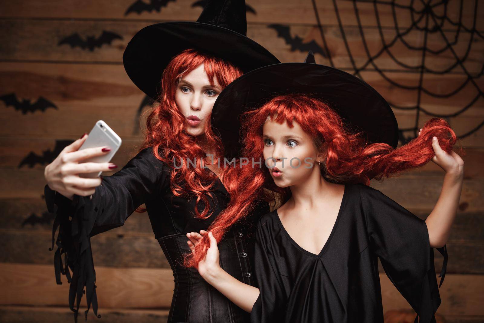 Halloween Concept - Beautiful caucasian mother and her daughter with long red hair in witch costumes taking a selfie with smartphone ready for celebrating Halloween