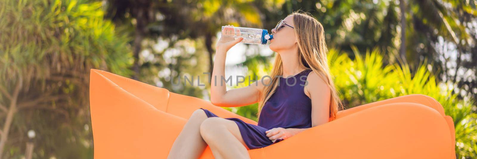BANNER, LONG FORMAT Summer lifestyle portrait of pretty girl sitting on the orange inflatable sofa and drinking water on the beach of tropical island. Relaxing and enjoying life on air bed by galitskaya