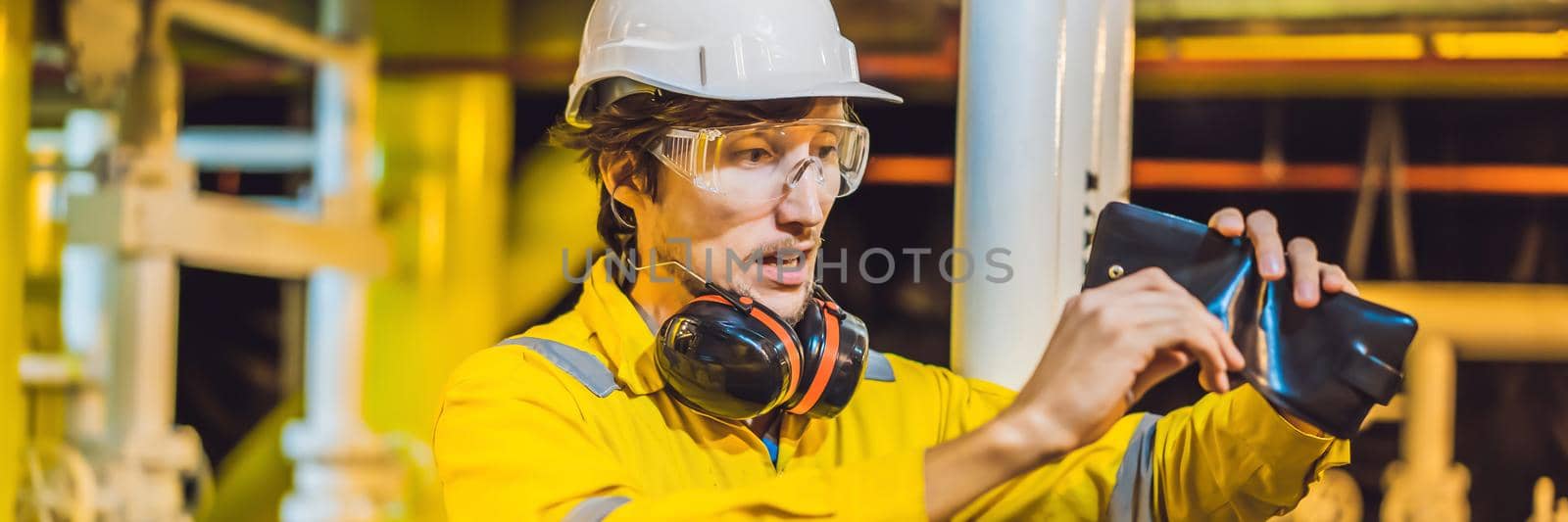 BANNER, LONG FORMAT Young man in a yellow work uniform, glasses and helmet in industrial environment,oil Platform or liquefied gas plant. looking into his empty wallet. Do not pay salary.