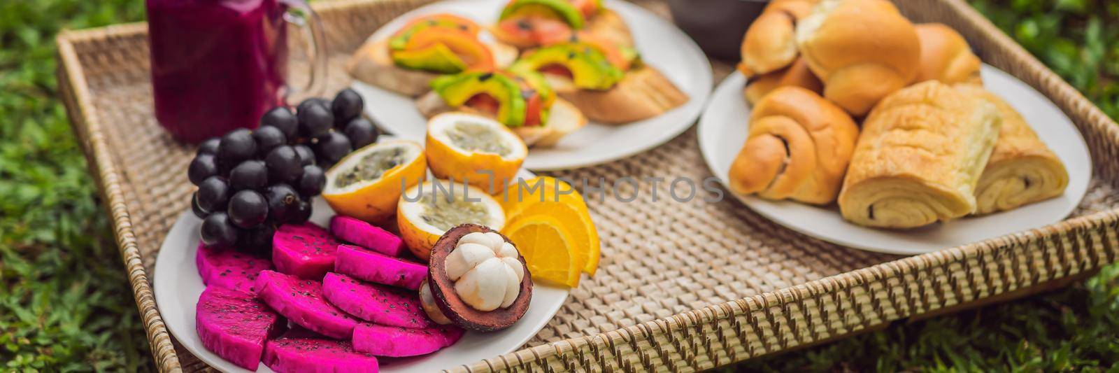 BANNER, LONG FORMAT Breakfast on a tray with fruit, buns, avocado sandwiches, smoothie bowl standing on the grass.