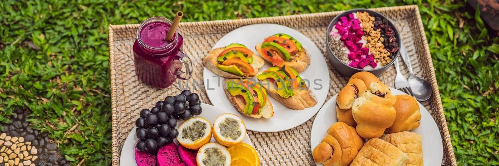 BANNER, LONG FORMAT Breakfast on a tray with fruit, buns, avocado sandwiches, smoothie bowl standing on the grass by galitskaya