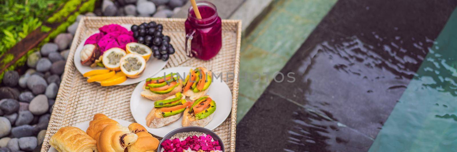 Breakfast on a tray with fruit, buns, avocado sandwiches, smoothie bowl by the pool. Summer healthy diet, vegan breakfast. Tasty vacation concept. BANNER, LONG FORMAT