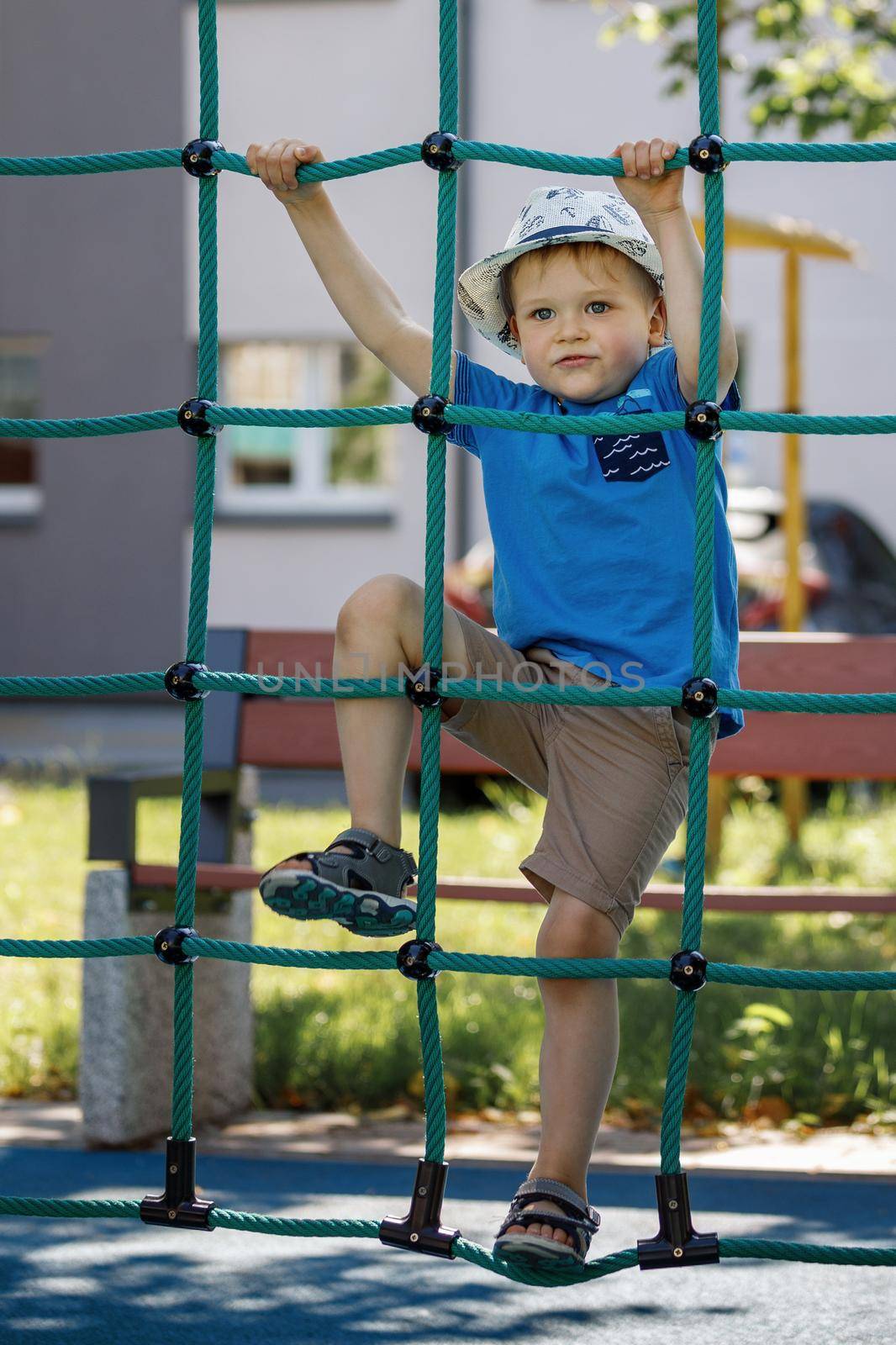 Portrait of cute smiling boy climbing up the rope net he look at camera. Concept of child development, sports and education.
