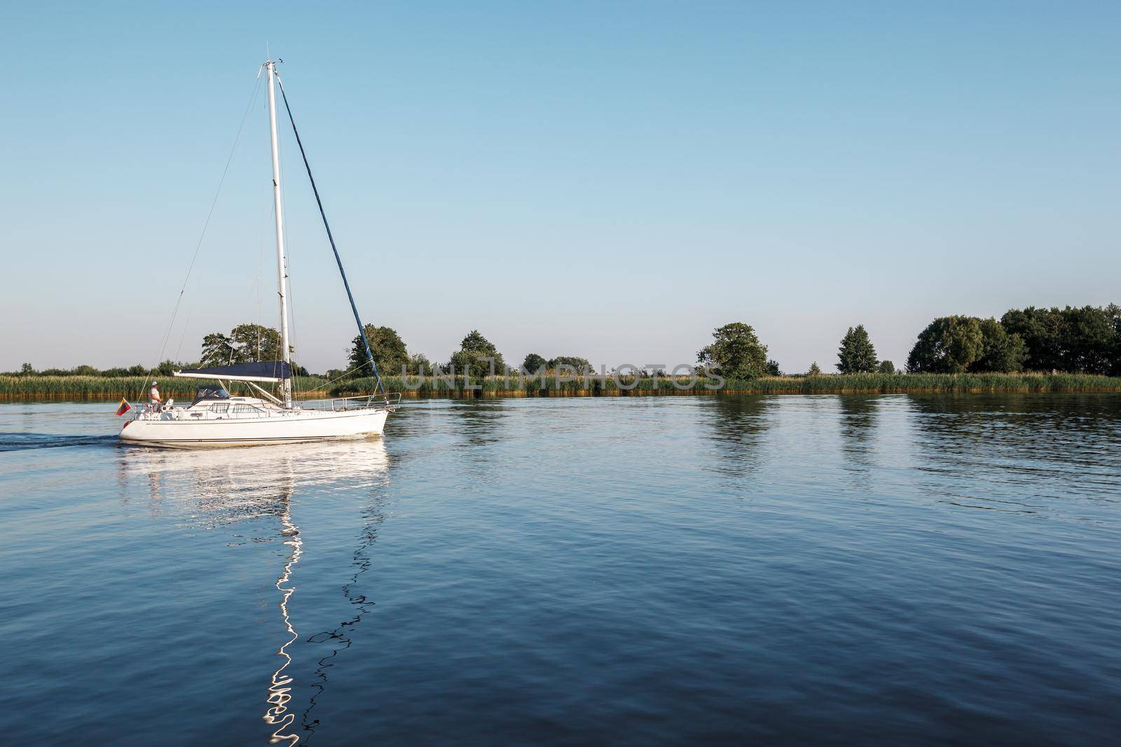 A white yacht with lowered sails sails in the lagoon. Quiet evening, well visible reflection of the ship's mast in the water. Horizontal photo. by Lincikas