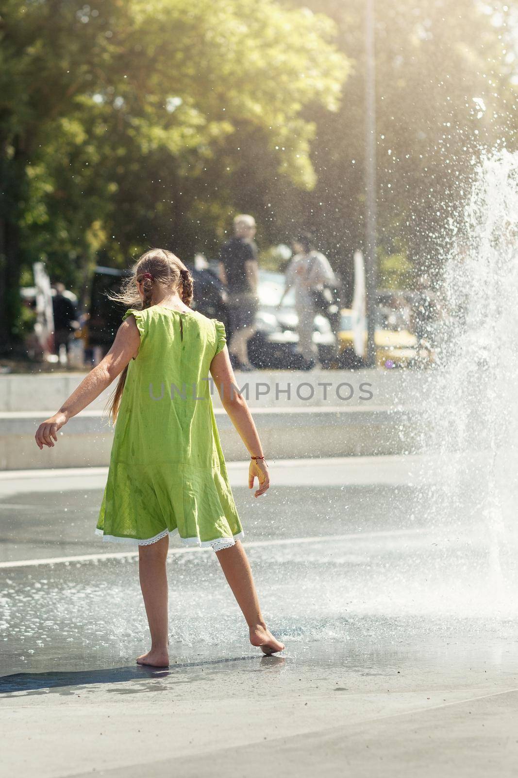 A young girl in a light green dress barefoot dances by a fountain in the city center.