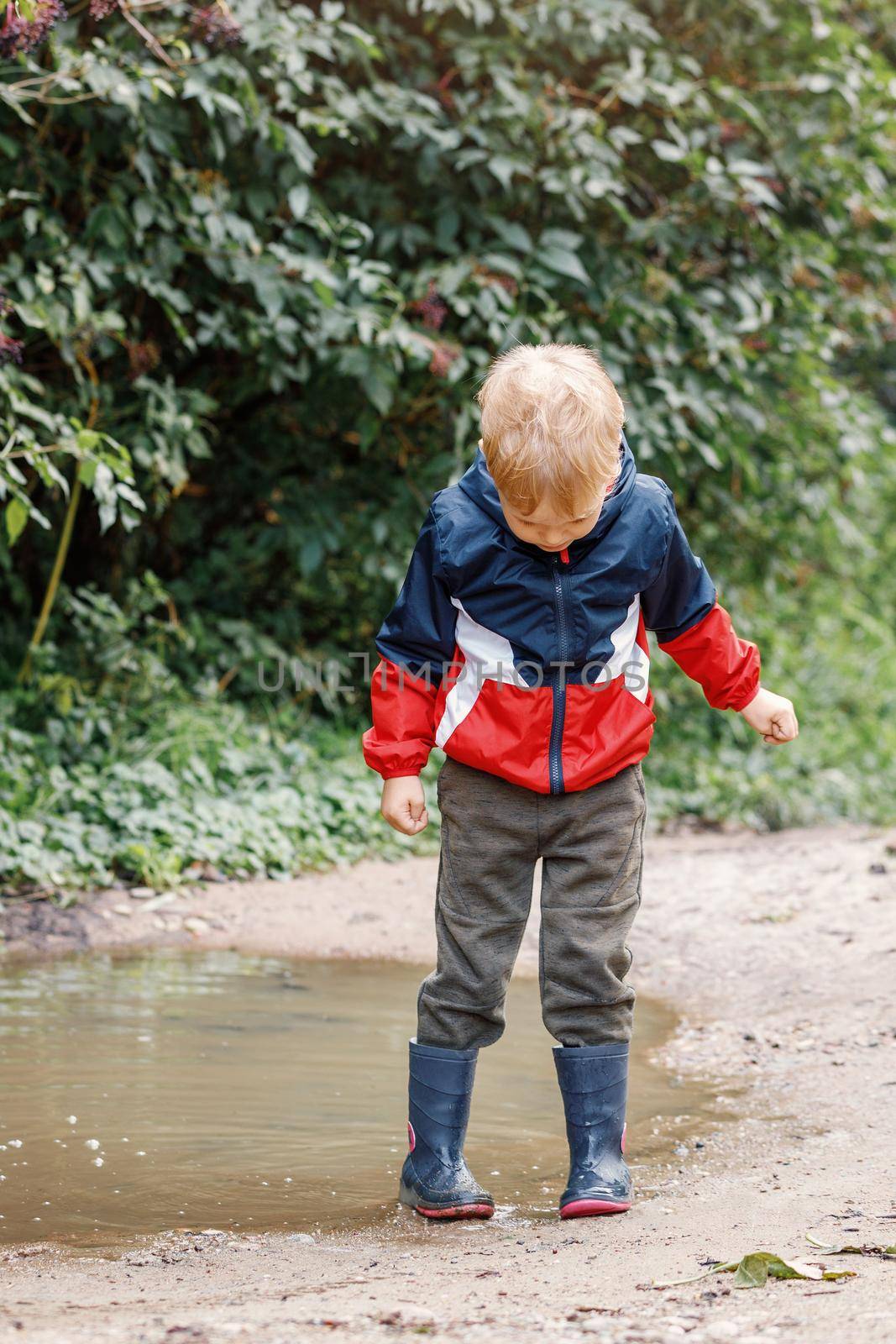 Child playing in puddle. Kids play outdoor by autumn in bad weather. Fall rainy weather outdoors activity for young children. Kid jumping in muddy puddles. Waterproof jacket and boots for little boy.