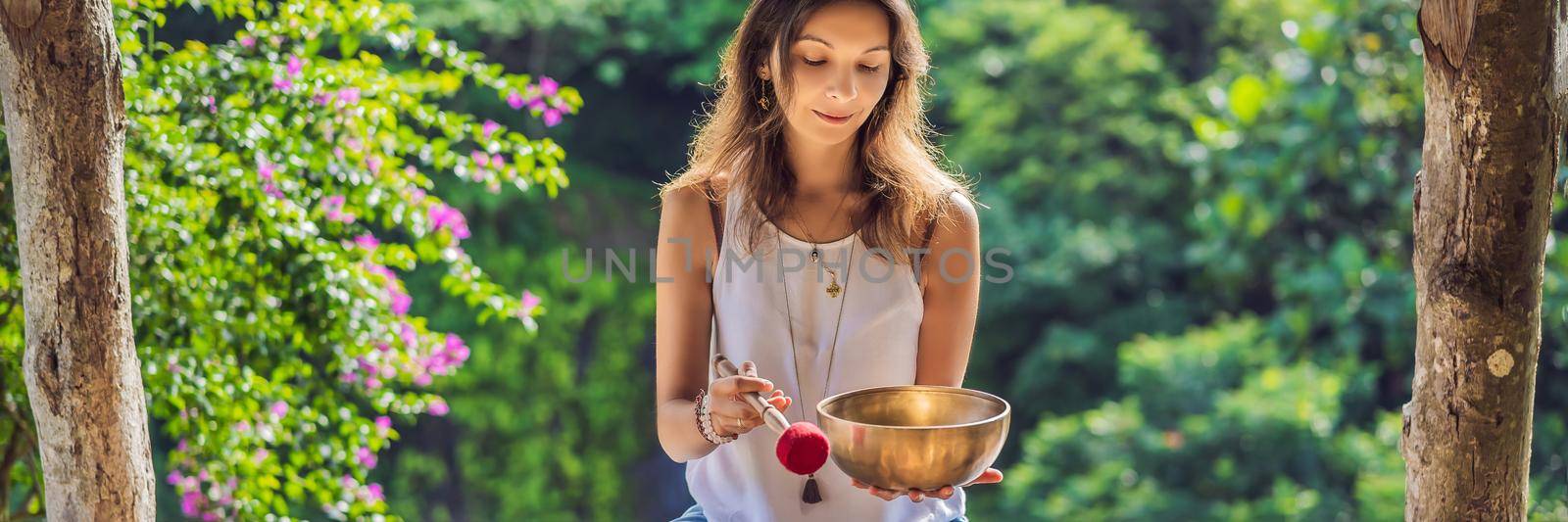 Woman playing on Tibetan singing bowl while sitting on yoga mat against a waterfall. Vintage tonned. Beautiful girl with mala beads meditating. BANNER, LONG FORMAT