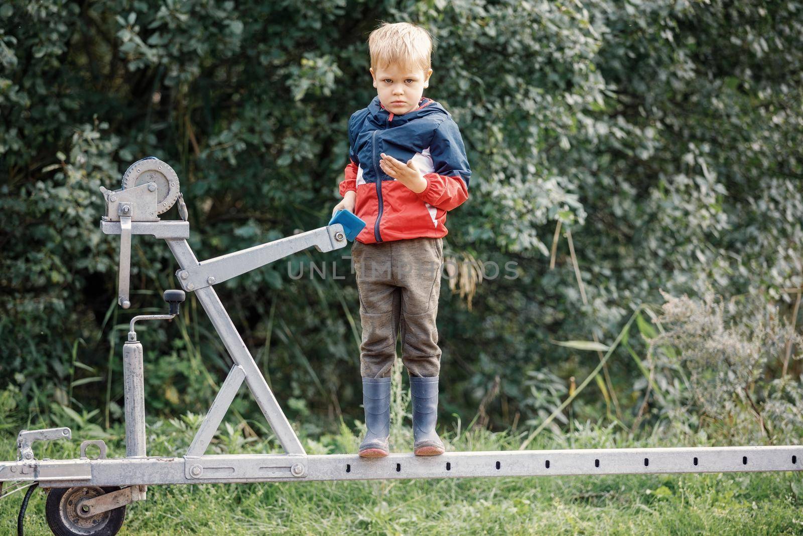 The blond boy with rubber boots climbs on boat trailer winch and poses for the camera against a background of green foliage.