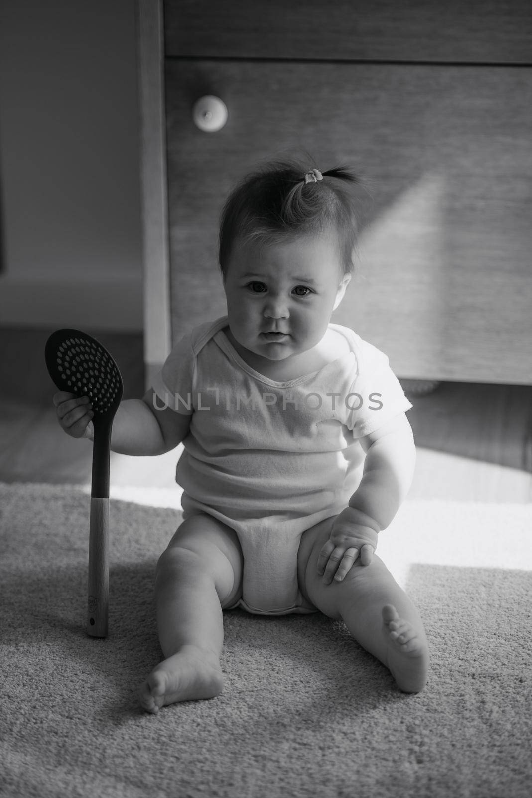 A black and white photo of a 7-month girl is sitting on the carpet dressed in a bodysuit at home. A cute infant is playing with a skimmer in the sunlight.
