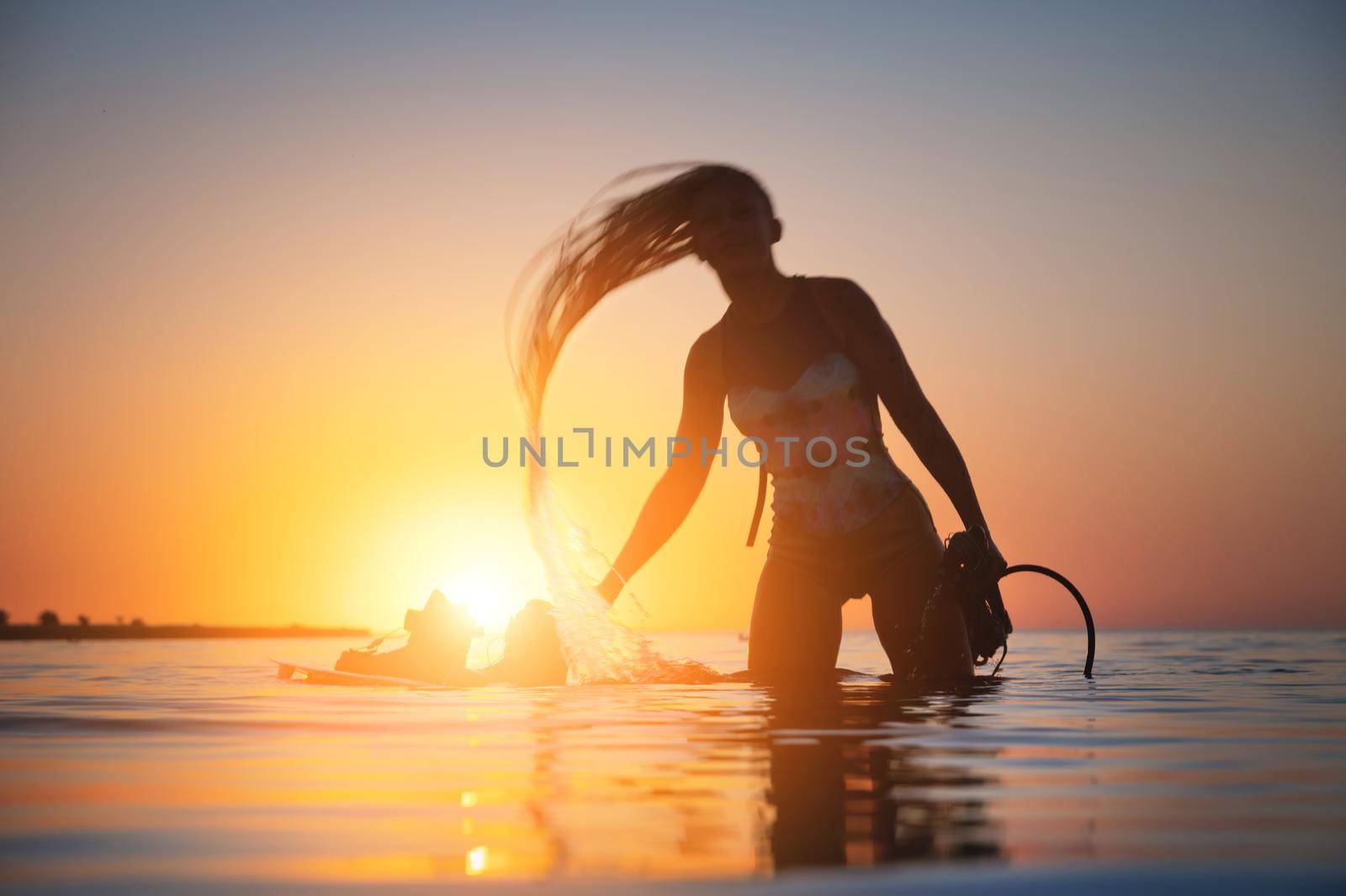 Silhouette frame. A young slim sexy female kitesurfer with a plank and a kiteboard stands in the water in the shallow water at sunset. Water sports. Stylized frame.