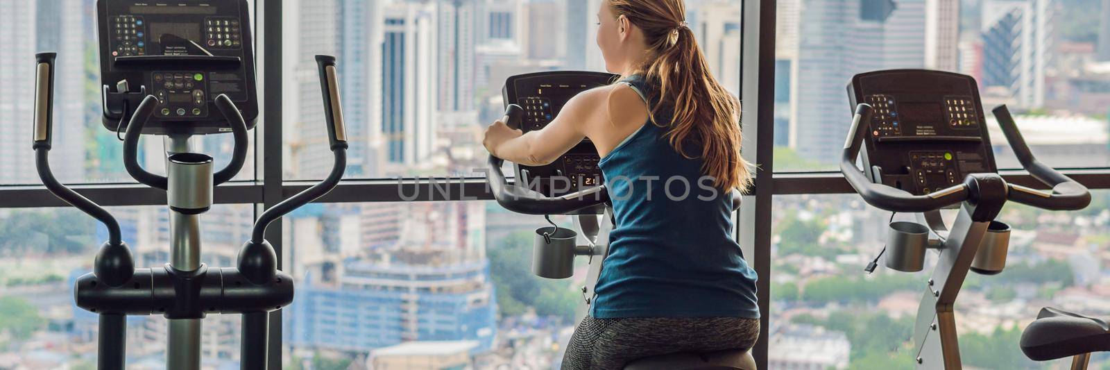 Young woman on a stationary bike in a gym on a big city background BANNER, LONG FORMAT by galitskaya