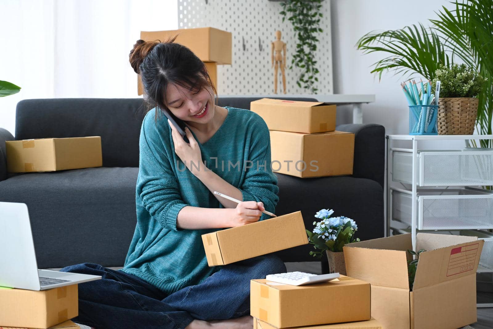Smiling asian woman online seller receive order on mobile phone and preparing parcel boxes of product for delivery.