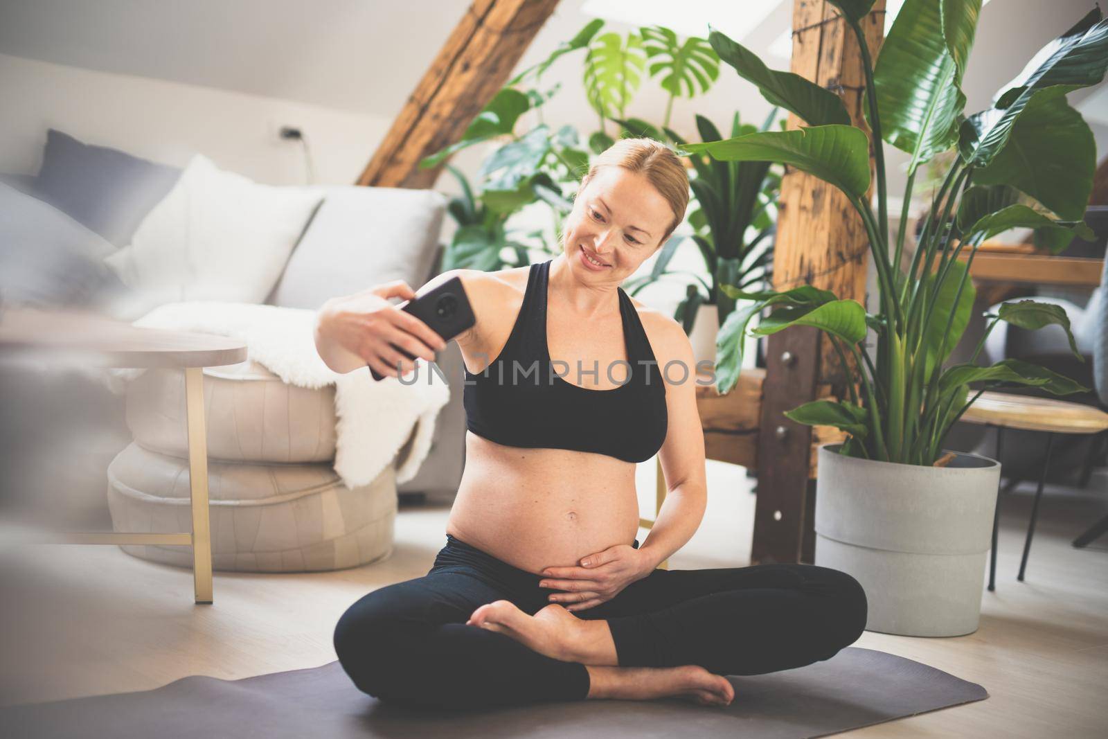 Young happy and cheerful beautiful pregnant woman taking selfie with her mobile phone while staying fit, sporty and active on her maternity leave. Motherhood, pregnancy, yoga concept
