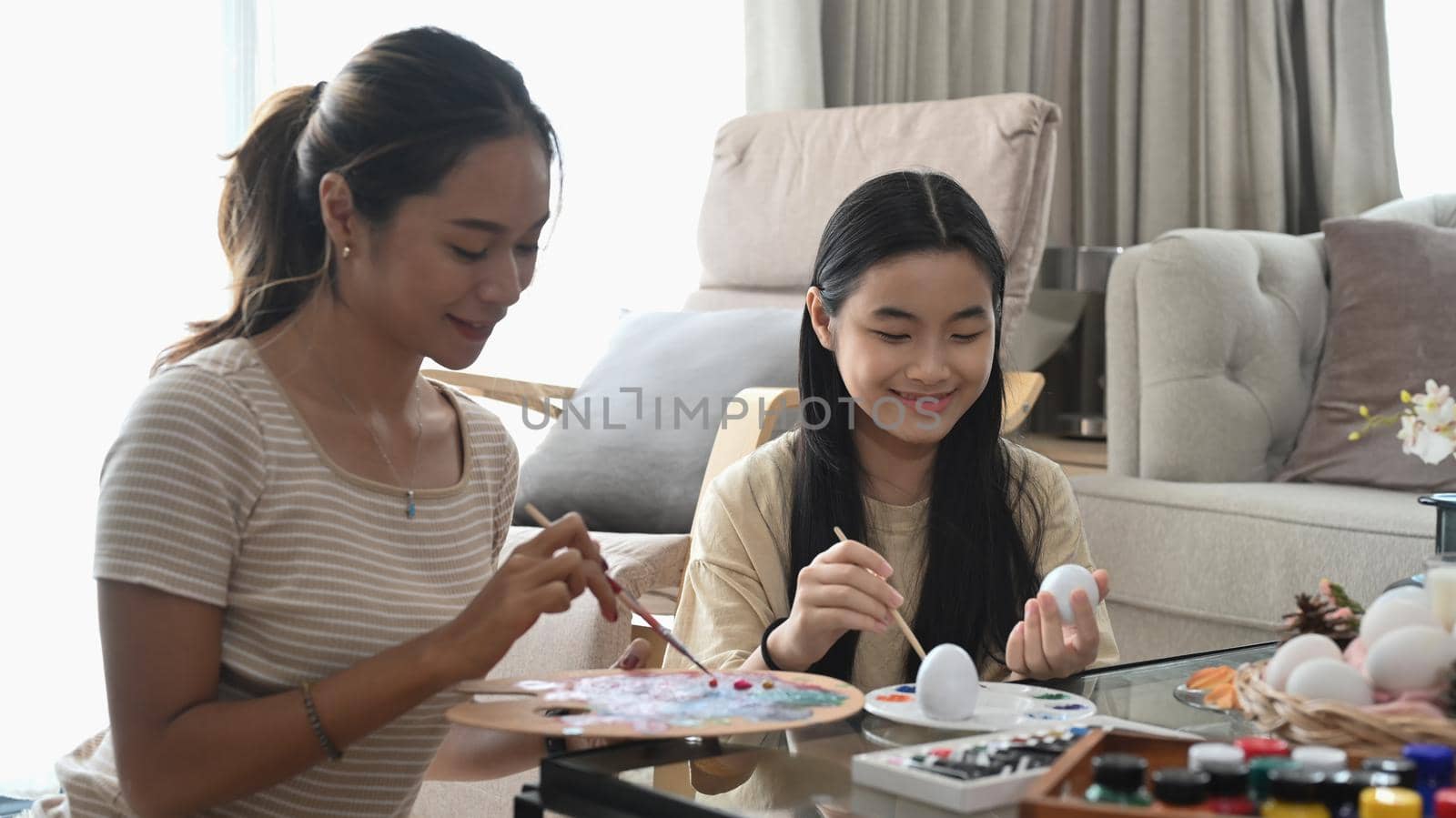Mom and daughter are sitting in bright living room and coloring Easter eggs. by prathanchorruangsak