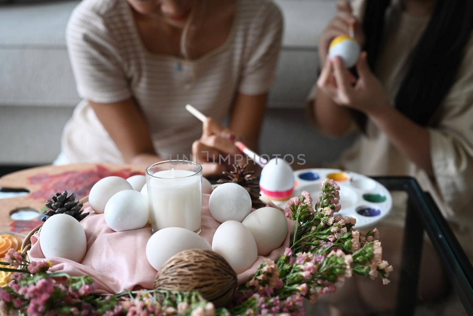 Easter basket with eggs, candle and spring flowers on table with mother and daughter painting Easter egg in background.